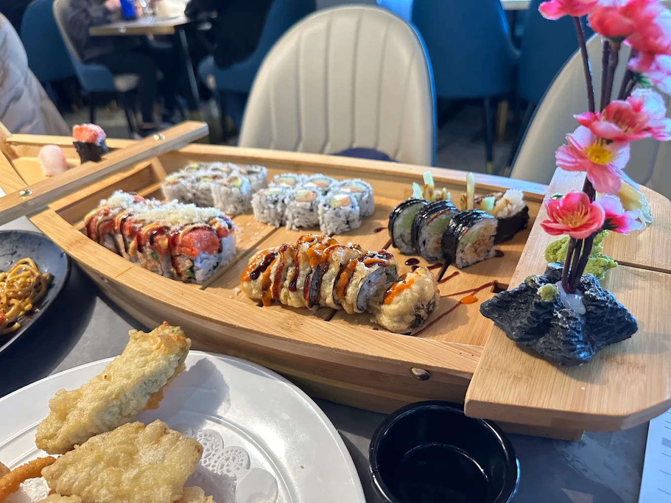 A wooden boat-shaped platter holds assorted sushi rolls, garnished with drizzles of sauce and accompanied by pink artificial flowers. A plate with tempura and a small bowl of soy sauce are nearby on the table.