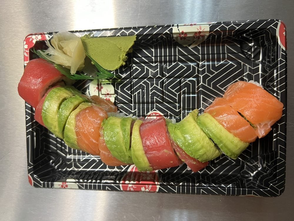 A sushi roll topped with slices of avocado, salmon, and tuna, arranged in a black plastic tray with wasabi and pickled ginger on the side.