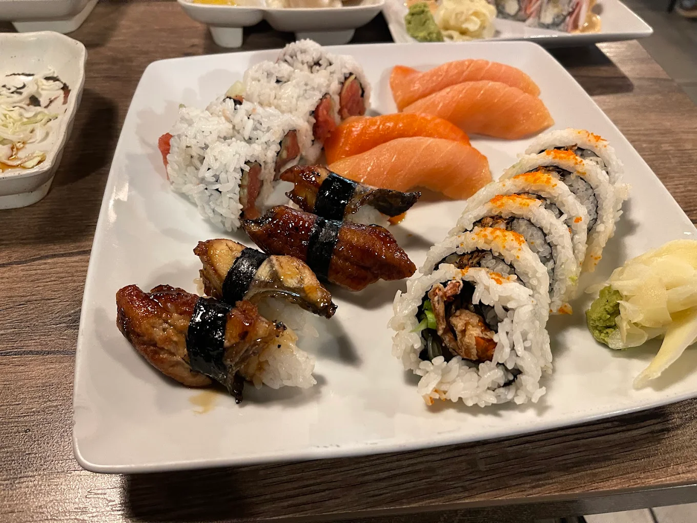 A plate of assorted sushi including nigiri with eel and salmon, sushi rolls with rice on the outside, wasabi, and pickled ginger on the side. The plate is on a wooden table with other dishes visible in the background.