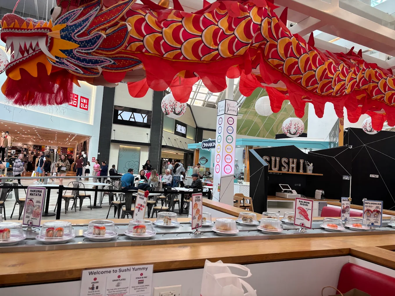 A sushi conveyor belt restaurant in a mall, with a large red dragon decoration overhead. Plates of sushi move along the belt, and diners are seated nearby. Several shops and people are visible in the background.