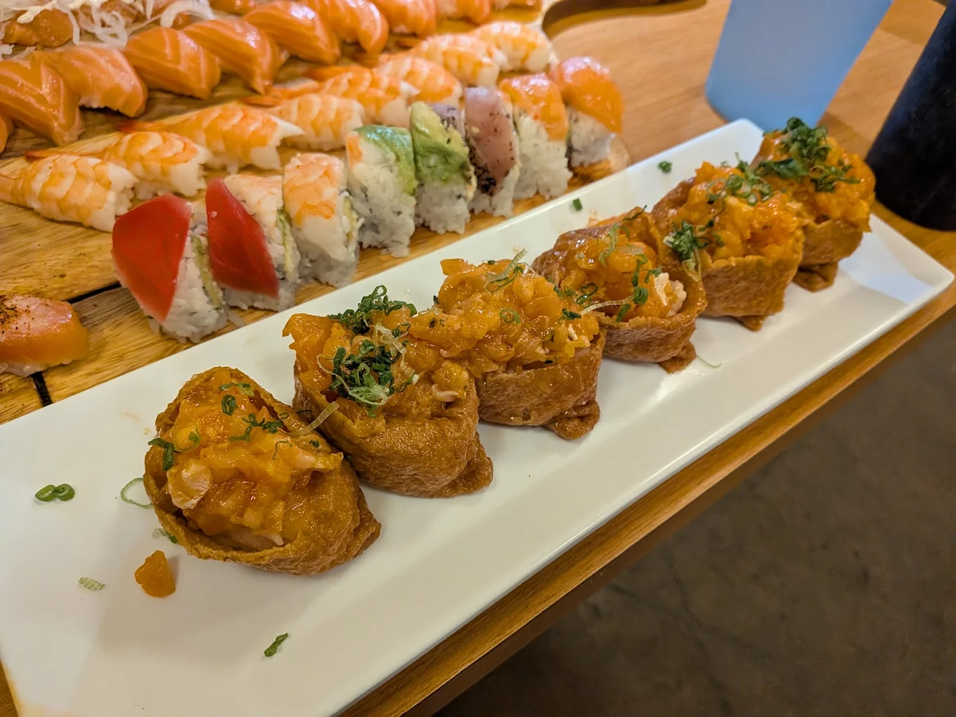 A white rectangular plate of six spicy sushi rolls topped with chopped green onions sits on a table, with assorted nigiri and maki sushi in the background.
