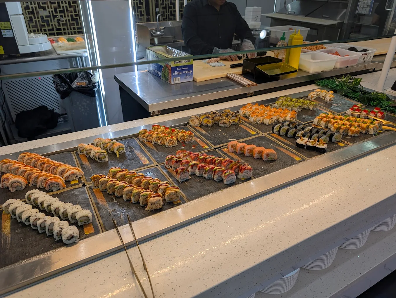 A variety of colorful sushi rolls are displayed on a counter at a sushi restaurant, arranged in neat rows behind a glass barrier, with utensils and condiments visible nearby.