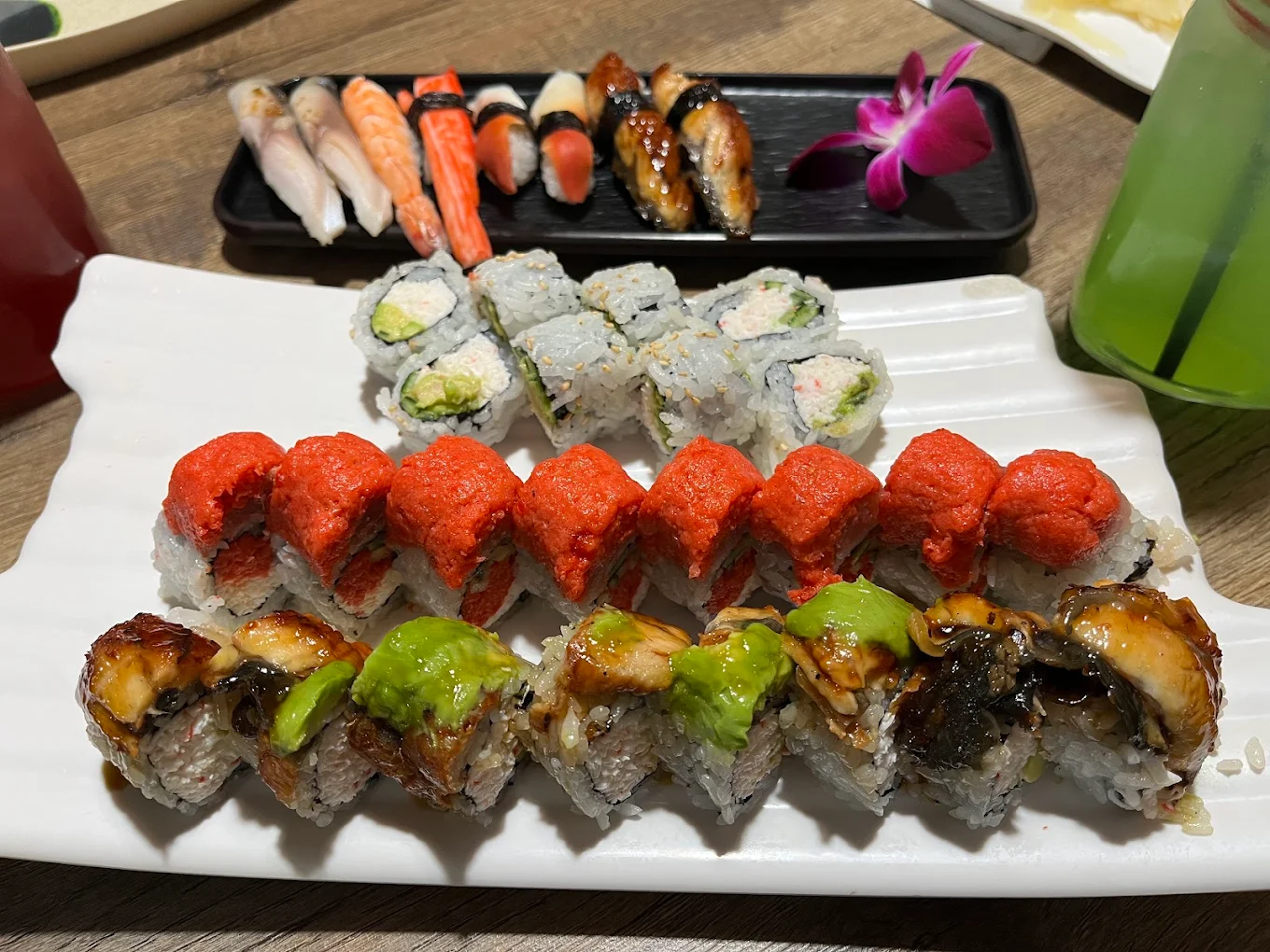 A plate of assorted sushi rolls with colorful toppings, including red, green, and brown sauces, sits in the foreground. In the background, there is a small tray of nigiri and a purple orchid garnish on a wooden table.