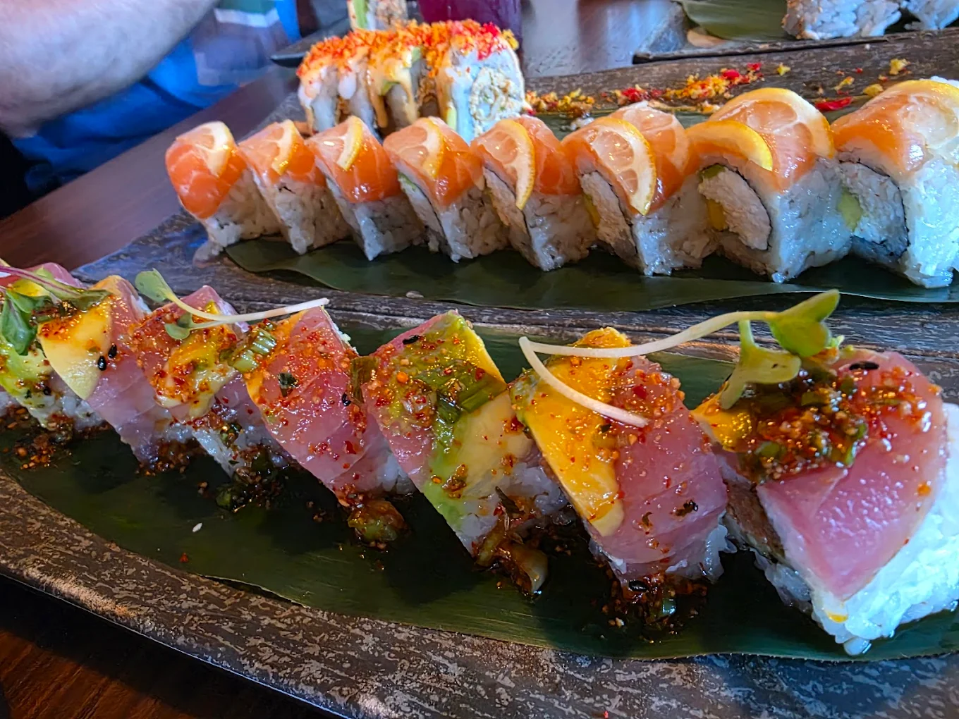 Close-up of two sushi rolls on a dark platter. The front roll is topped with raw tuna, avocado, and microgreens, garnished with sauce and spices. The back roll features salmon and lemon slices on top.