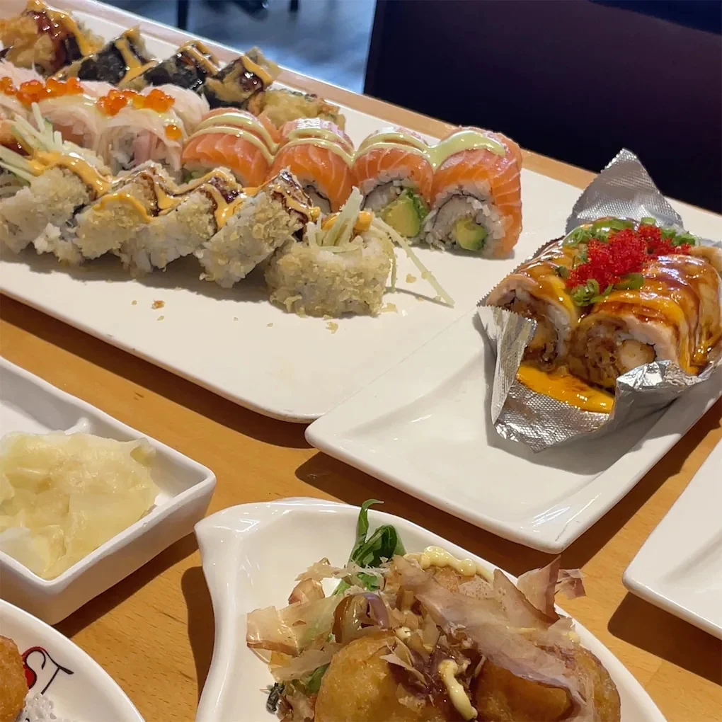 A variety of sushi rolls with colorful toppings, garnishes, and sauces are arranged on white plates on a wooden table, along with small side dishes of ginger and fried appetizers.