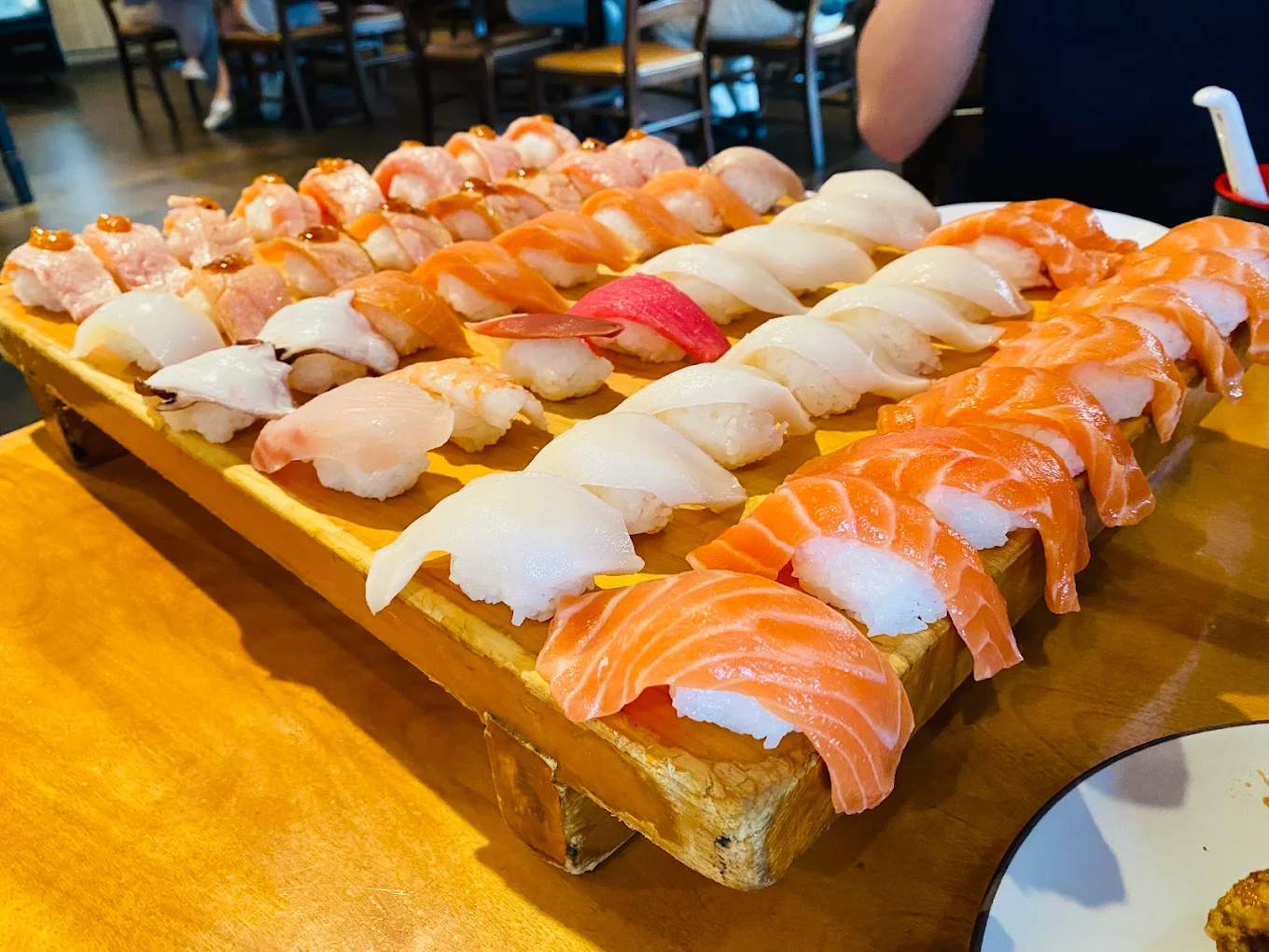 A large wooden tray filled with assorted nigiri sushi, including various types of fish such as salmon and white fish, arranged neatly on a restaurant table. Diners are seen sitting in the background.