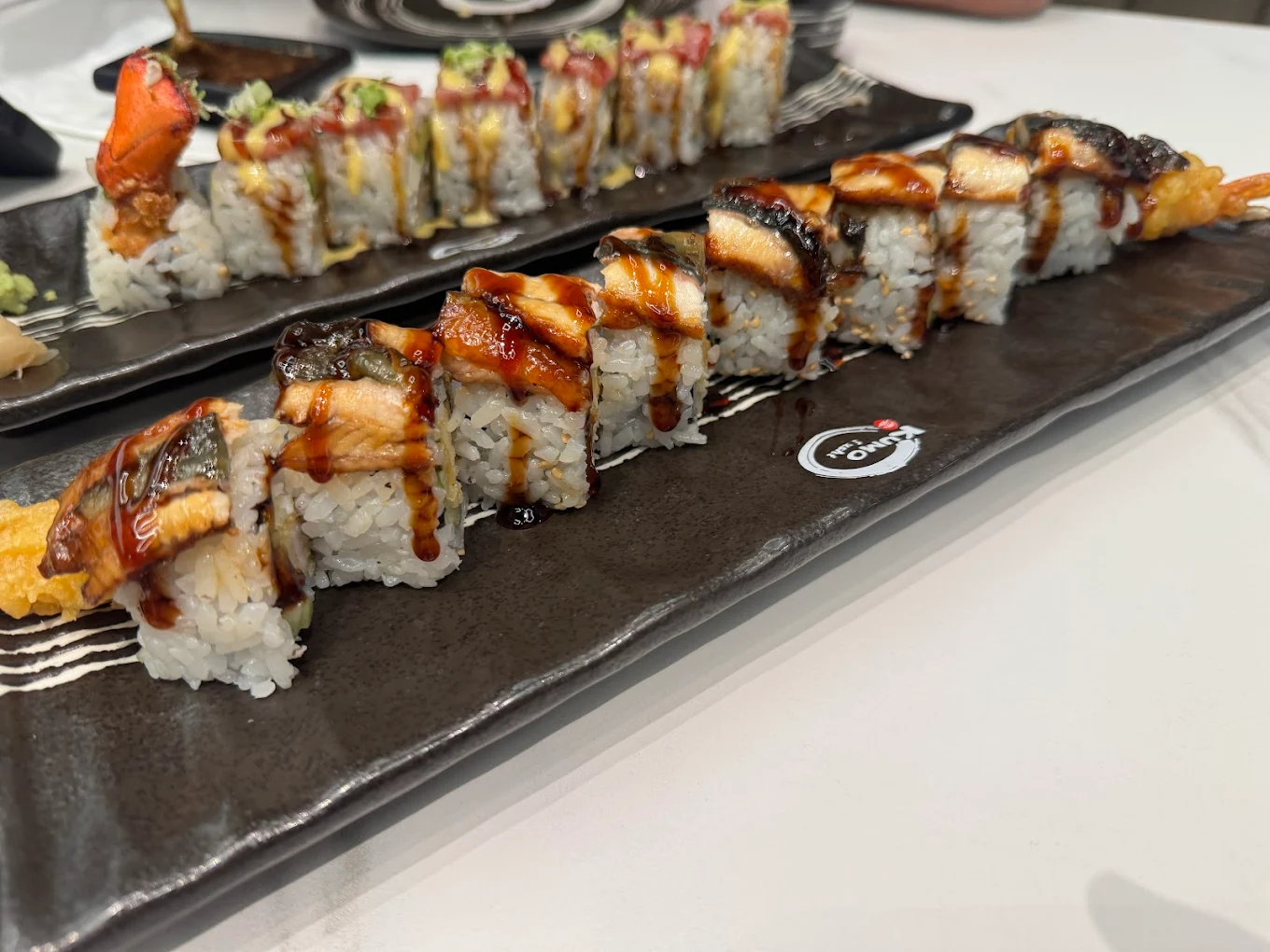 A close-up of two plates of sushi rolls on a table. The front plate features rolls topped with eel and sauce, while the plate in the background has assorted sushi rolls with colorful toppings.