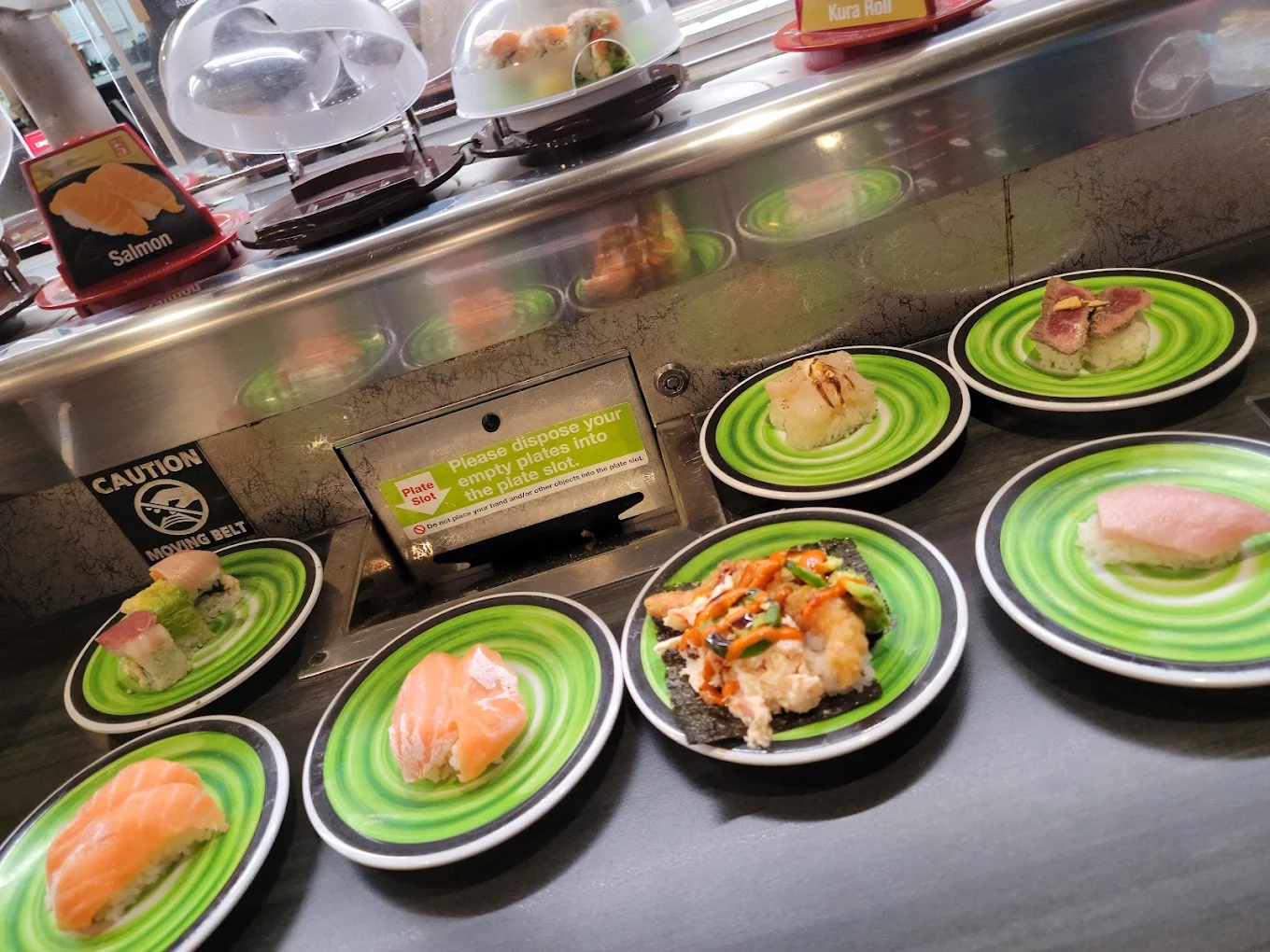 Plates of assorted sushi on a green conveyor belt at a sushi restaurant, with a sign asking customers to deposit empty plates into a slot. Various sushi types are visible on the plates.