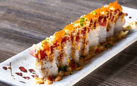 A rectangular sushi roll cut into pieces, topped with orange roe, spicy mayo, green onions, crispy flakes, and sauce, arranged on a long white plate on a wooden table.
