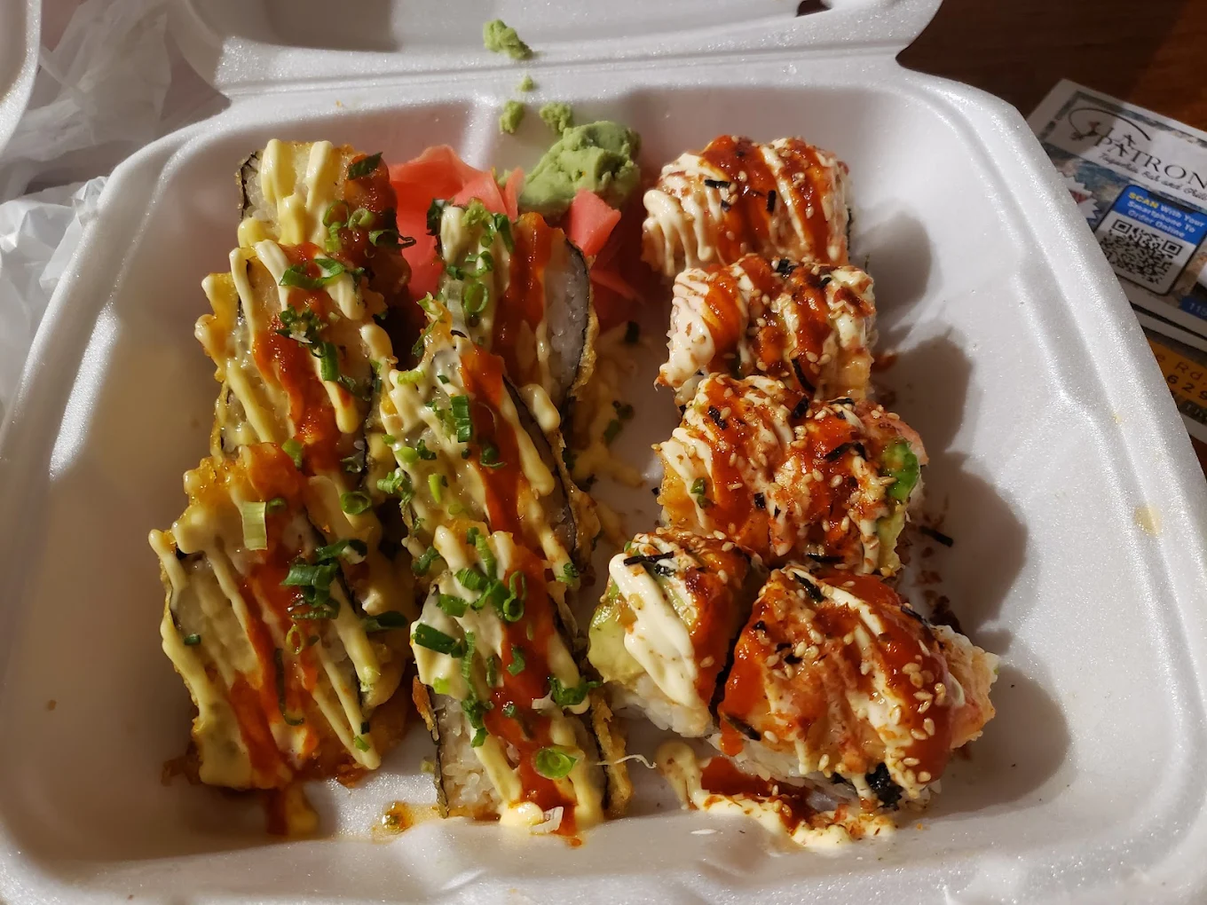 Two types of sushi rolls in a white takeout container, topped with drizzled sauces, sesame seeds, green onions, and accompanied by pickled ginger and wasabi.