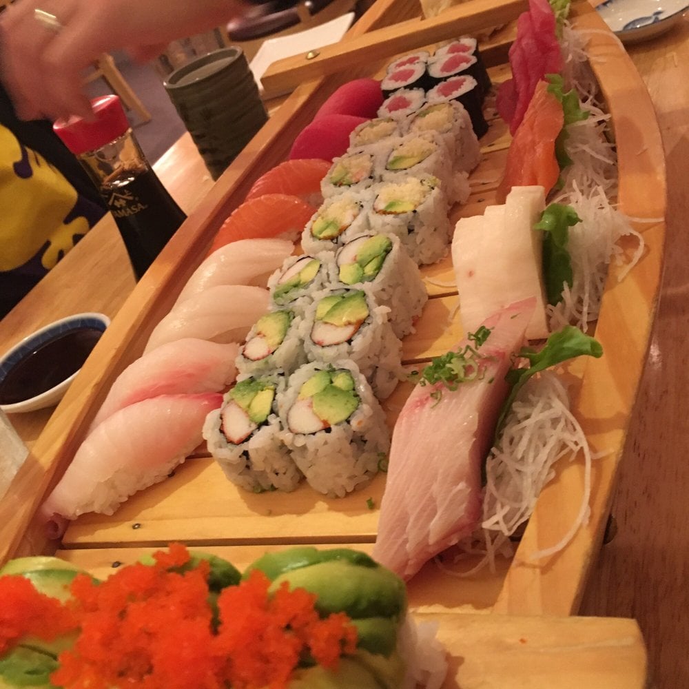A wooden tray filled with assorted sushi, including nigiri, sashimi, and rolls with avocado, fish, and vegetables. A hand reaches for soy sauce in the background.