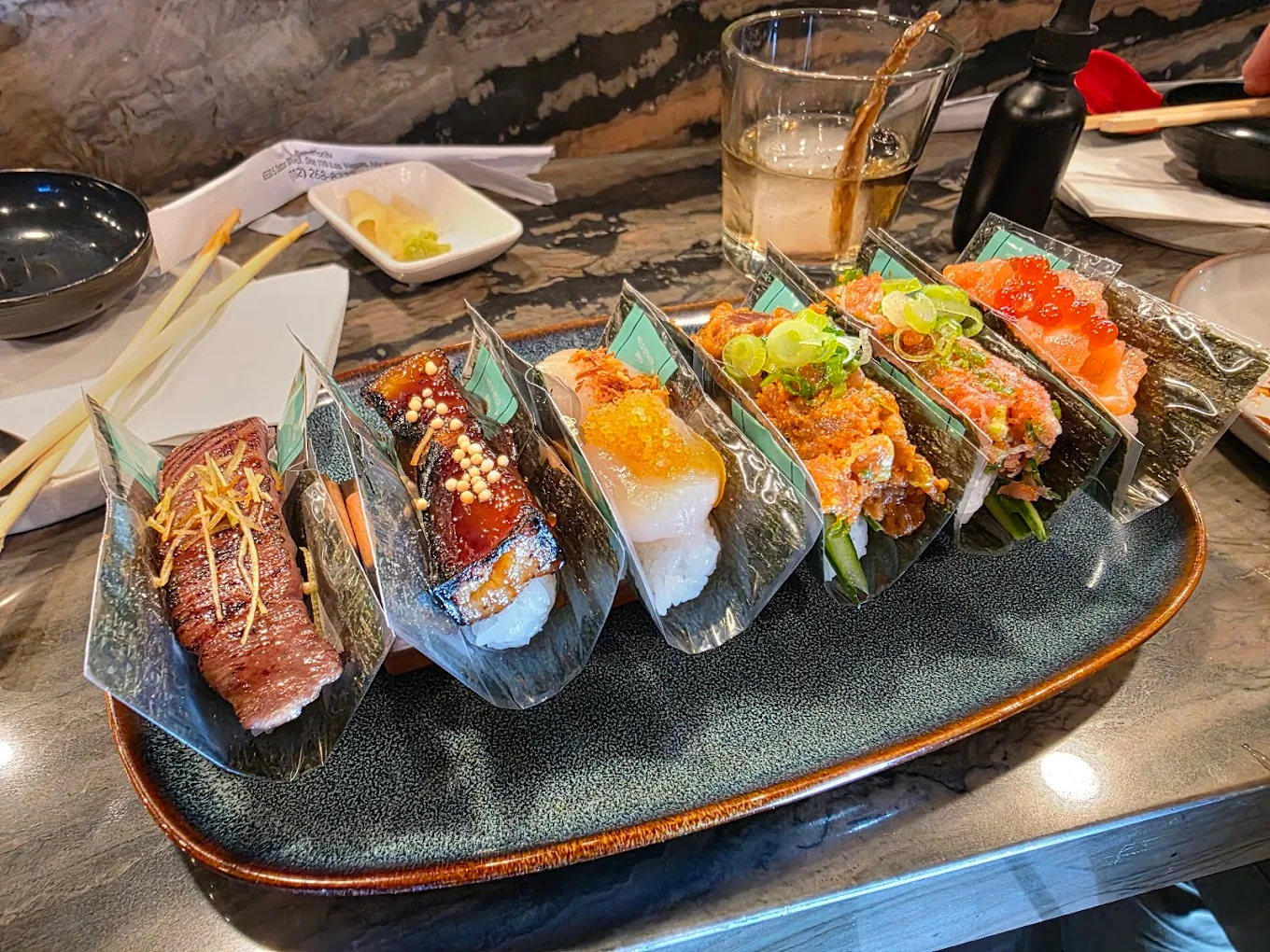 A plate of six assorted sushi hand rolls, each wrapped in seaweed and filled with different ingredients, is displayed on a restaurant table with chopsticks, sauce, wasabi, and a drink in the background.