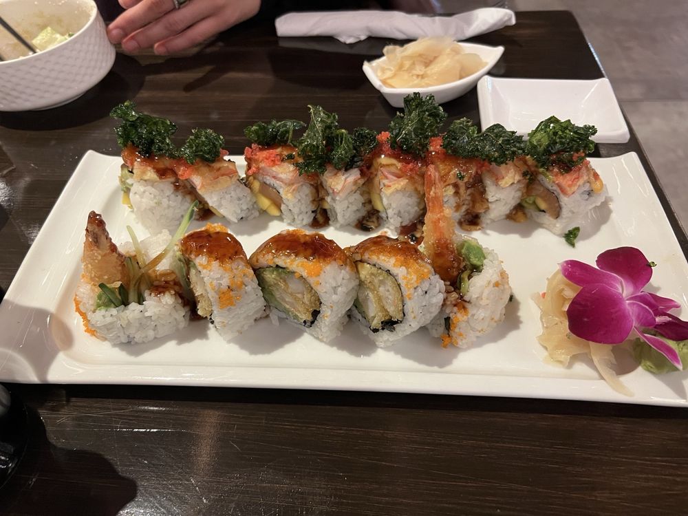 A rectangular white plate with assorted sushi rolls topped with greens and sauce, garnished with a purple orchid and pickled ginger, on a dark table. A hand, bowl, and small dish are visible in the background.