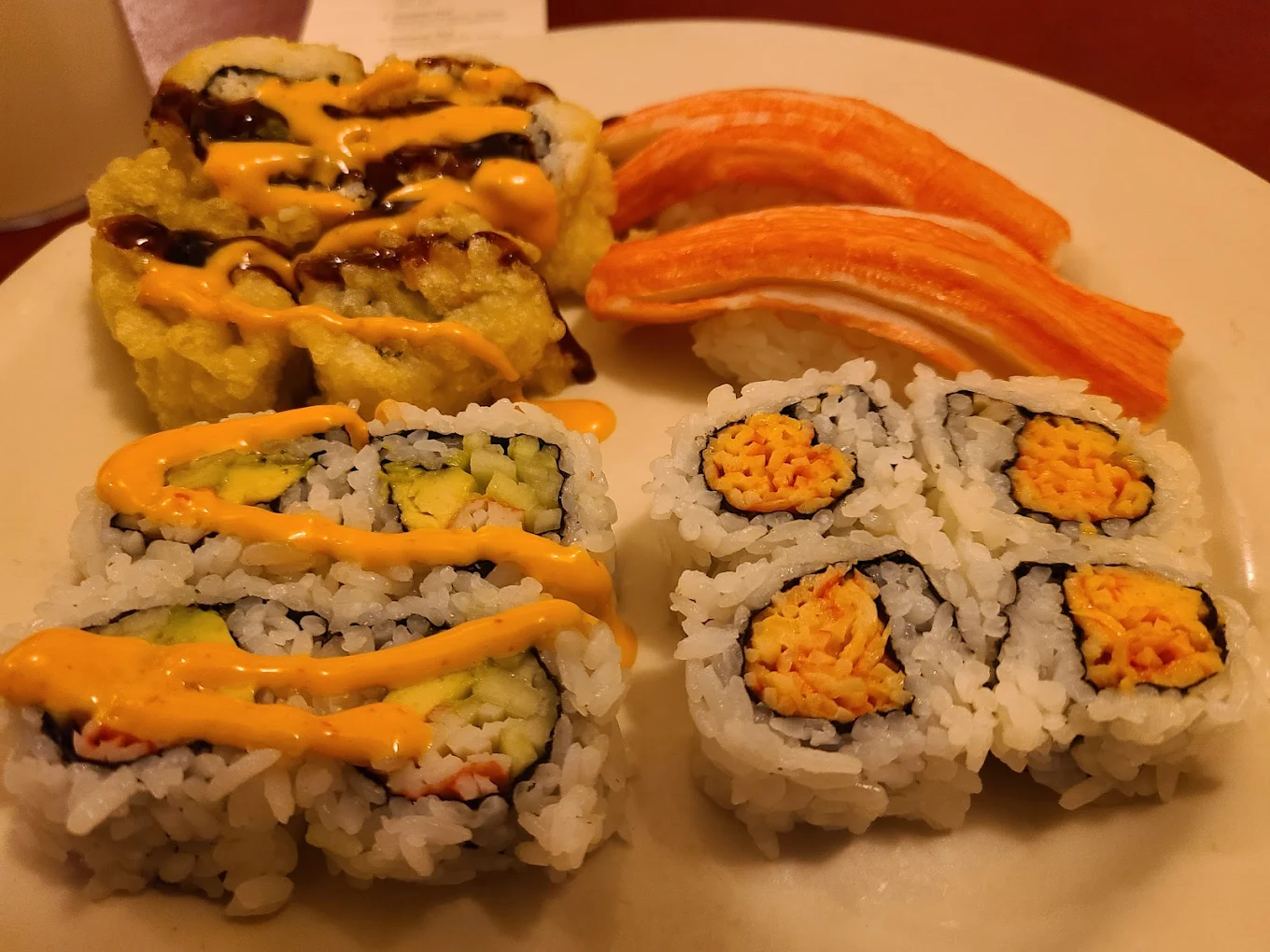 A plate with four sushi varieties: two pieces with orange sauce, two pieces of nigiri with imitation crab, four California rolls, and four spicy rolls topped with orange sauce.