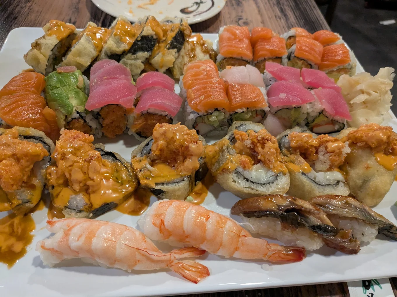 A plate filled with assorted sushi rolls, including pieces topped with salmon, tuna, shrimp, and eel, some garnished with spicy sauce, and accompanied by ginger slices on the side.