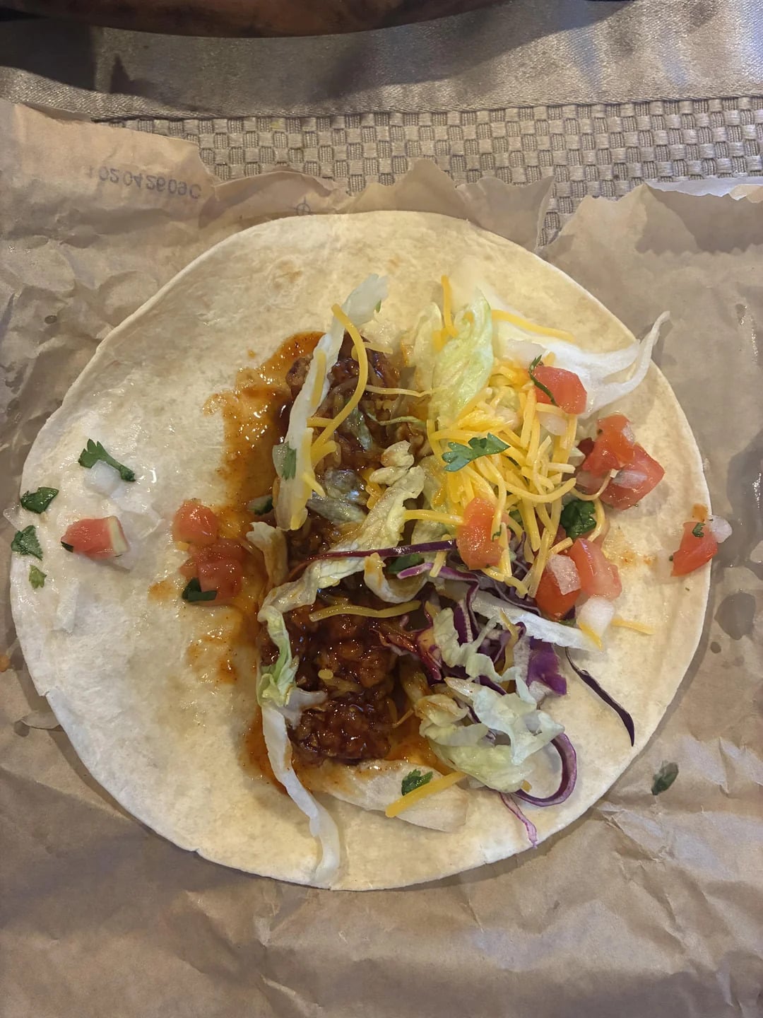A soft tortilla topped with seasoned ground beef, shredded lettuce, shredded cheese, chopped tomatoes, and cilantro, on brown paper.