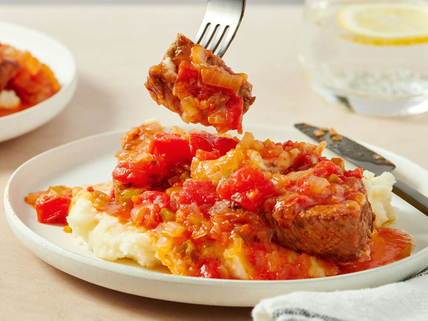 A plate of mashed potatoes topped with beef stew in tomato sauce; a fork holds a bite-sized piece above the plate. A glass of water with lemon is in the background.