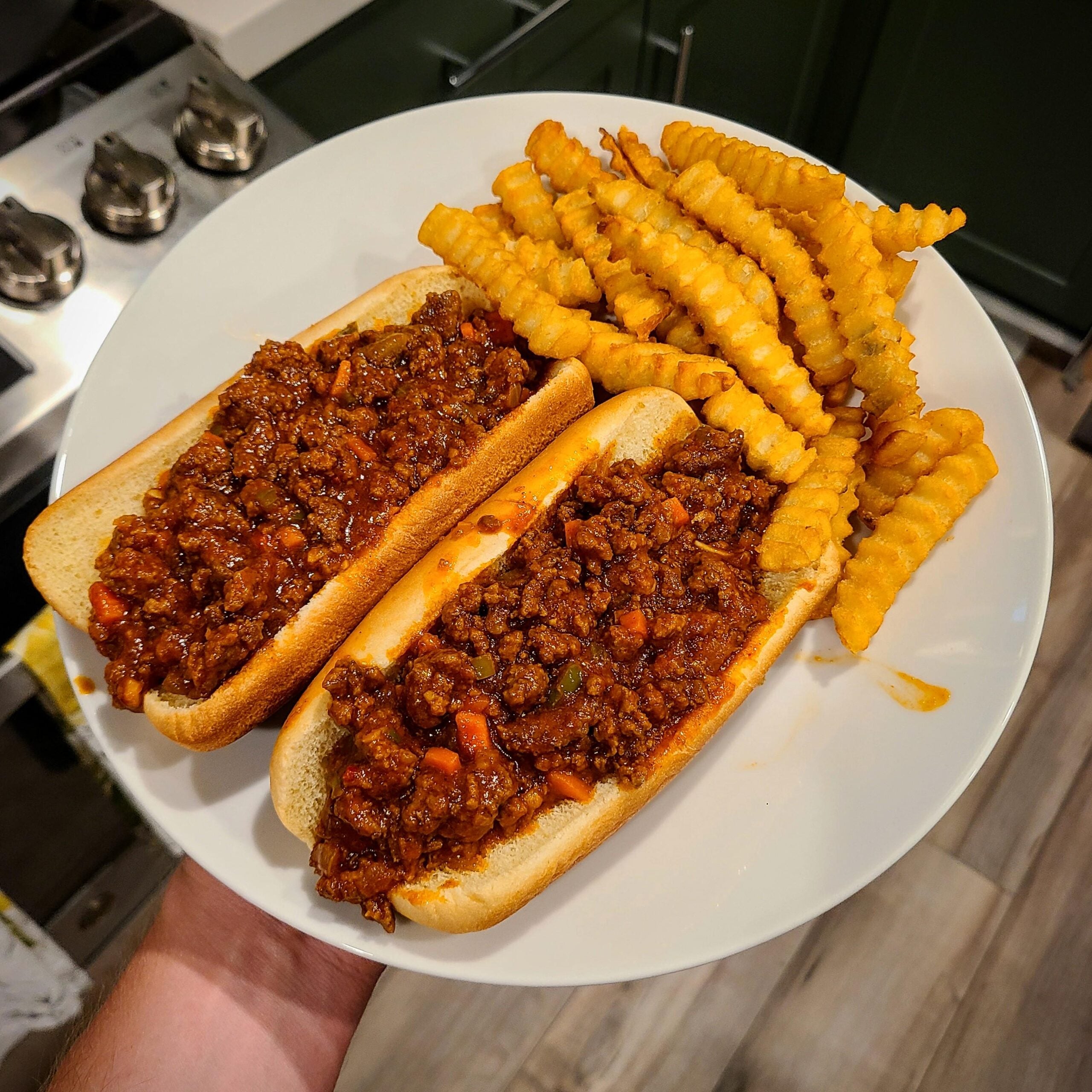 A plate with two sloppy joes in buns and a serving of crinkle-cut French fries, held above a kitchen floor near a stove and green cabinets.