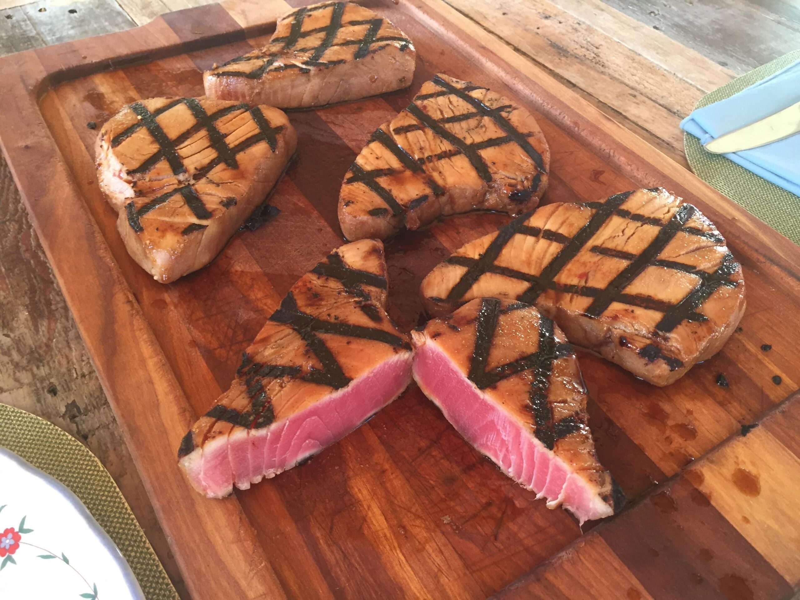 Five grilled tuna steaks with visible grill marks on a wooden cutting board; two steaks are cut to reveal a pink, rare center. The background includes a table setting with utensils and a green placemat.