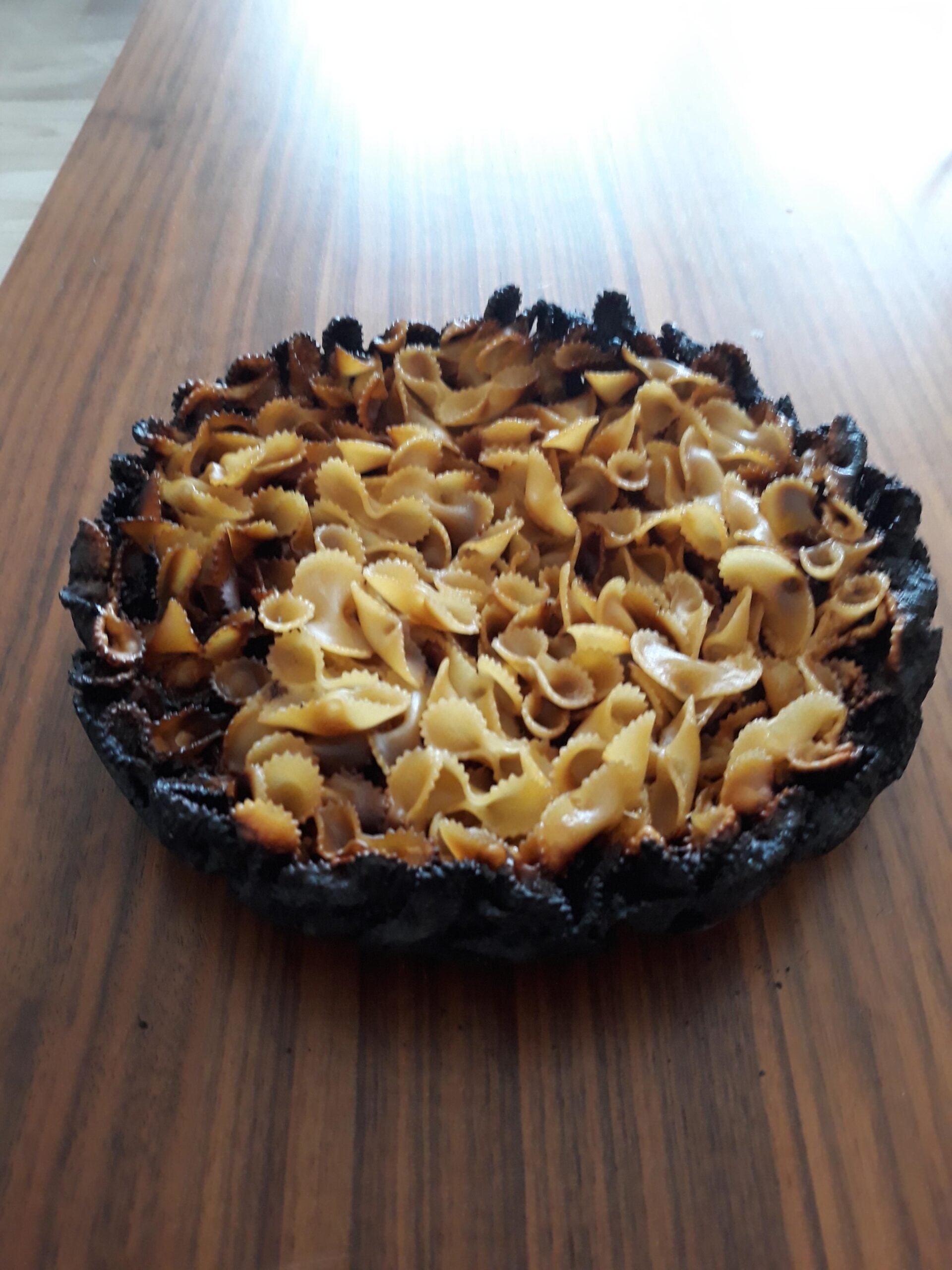 A burnt pie crust filled with bowtie pasta sits on a wooden table. The edges of the crust are blackened, and the pasta appears unevenly cooked.