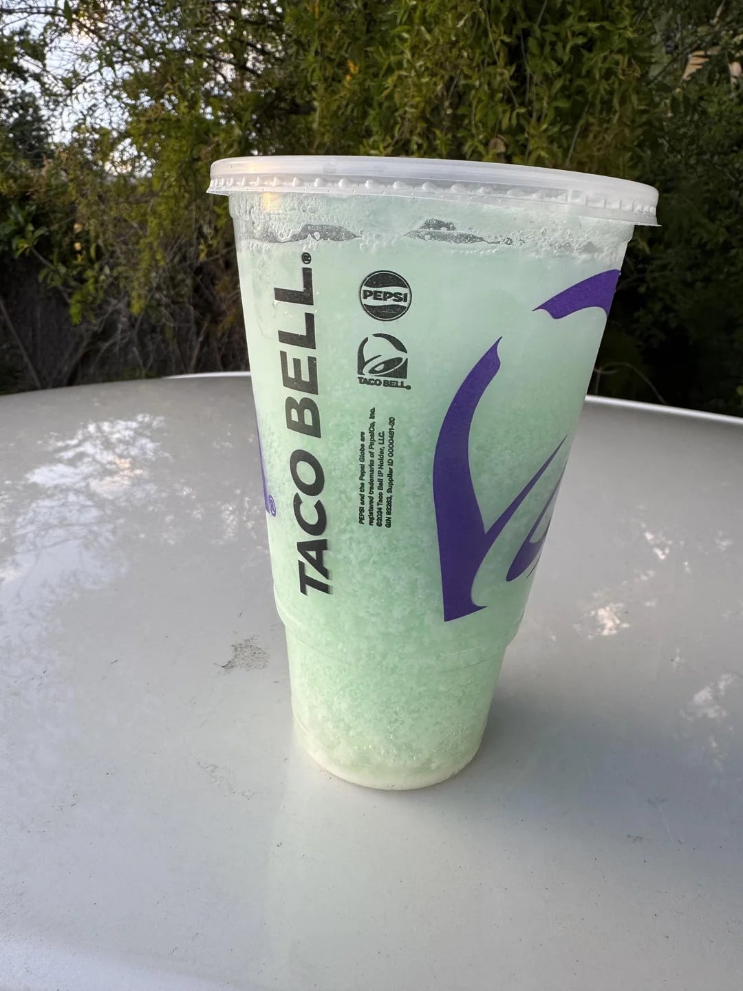 A large Taco Bell cup filled with a light green frozen drink sits on a white surface outdoors, with trees and greenery in the background.
