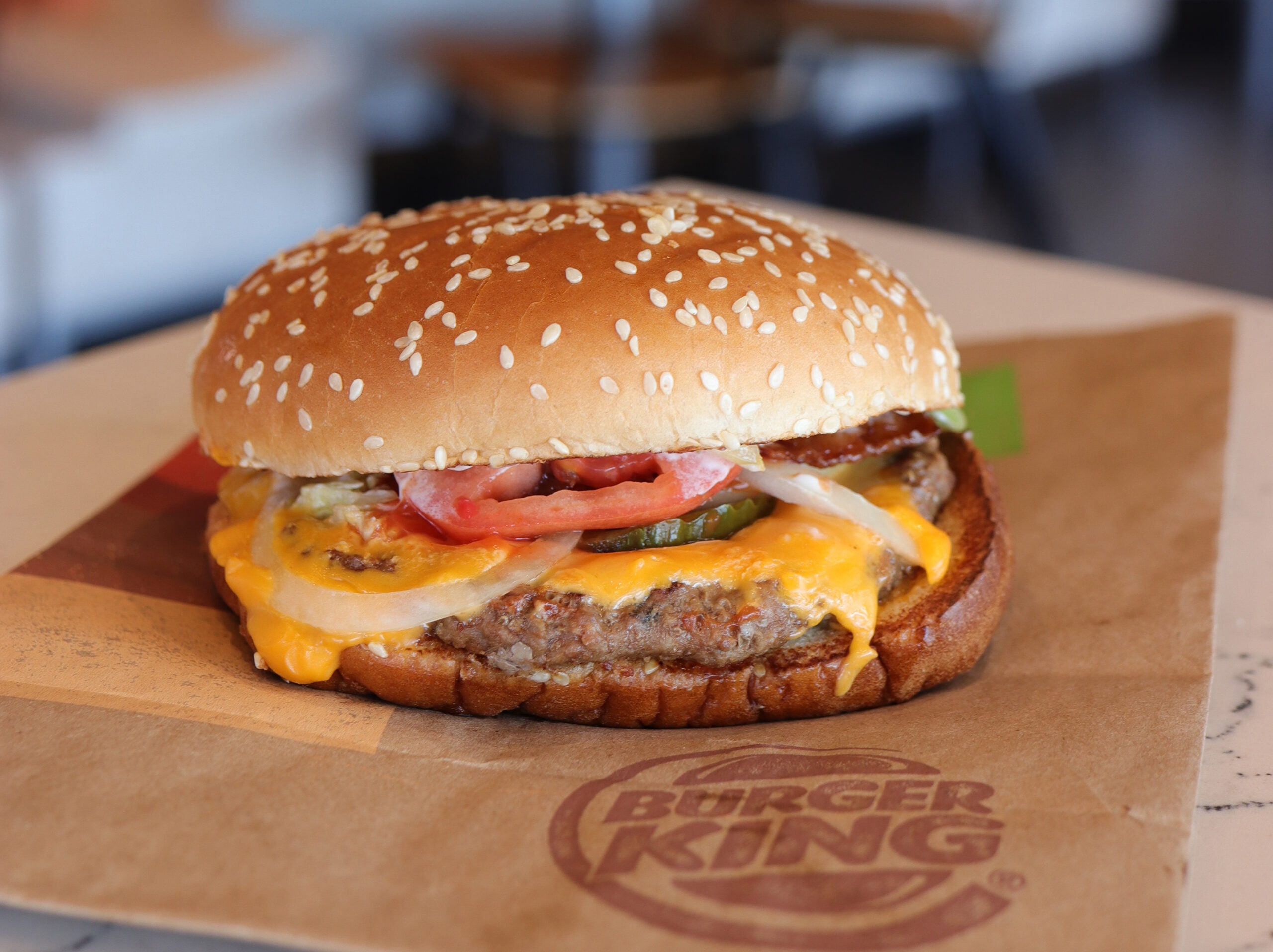 A Burger King cheeseburger with melted cheese, tomato, pickles, and onions on a sesame seed bun, placed on a Burger King branded wrapper.