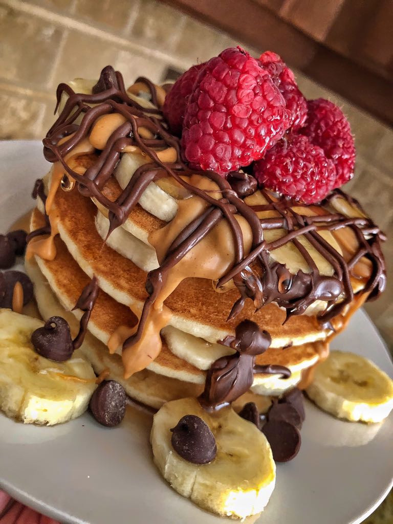 A stack of pancakes topped with raspberries, drizzled with chocolate and peanut butter, and garnished with banana slices and chocolate chips.