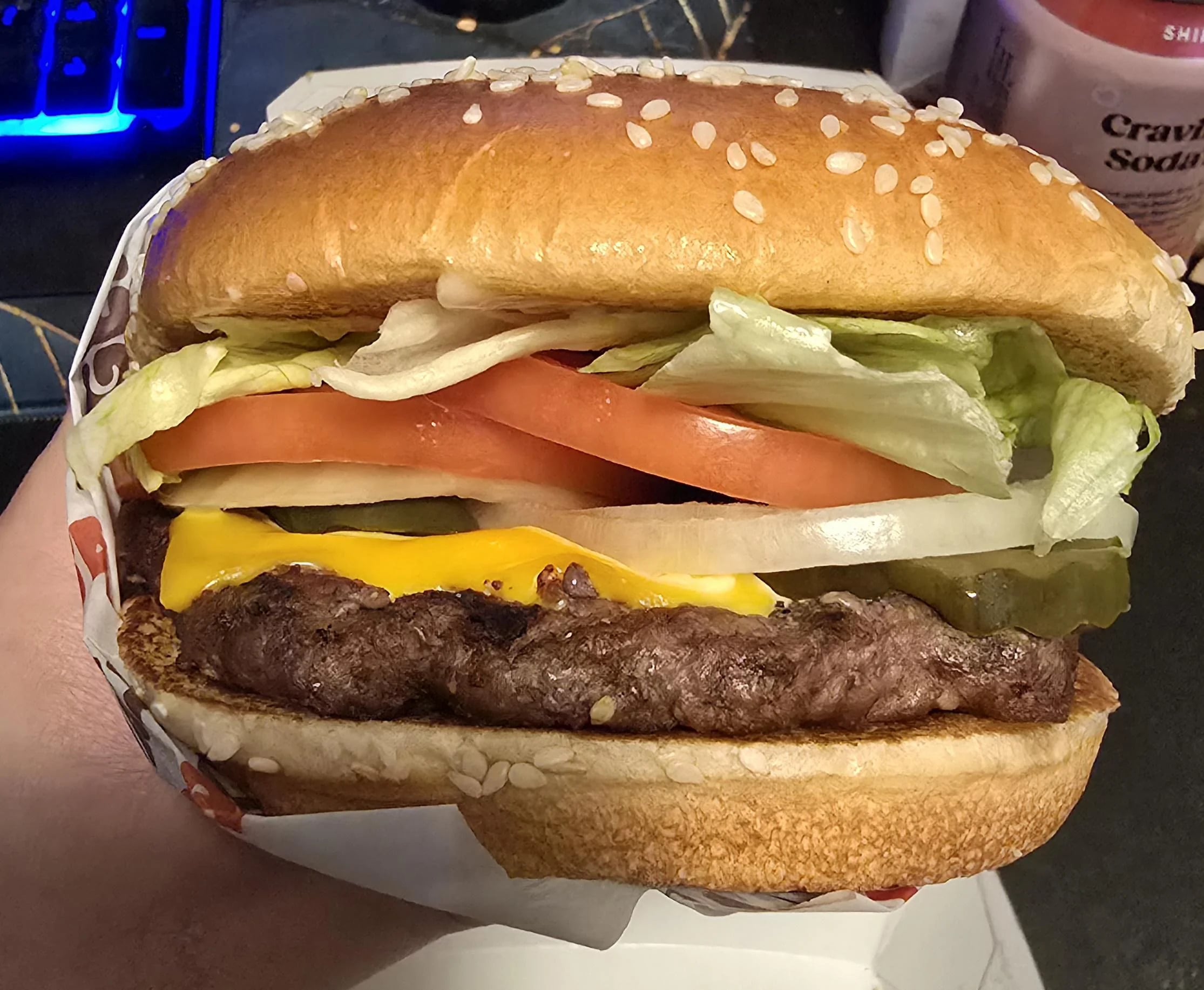 A hand holds a cheeseburger with a sesame seed bun, lettuce, tomato, onion, pickles, cheddar cheese, and a beef patty. A drink cup and part of a keyboard are visible in the background.
