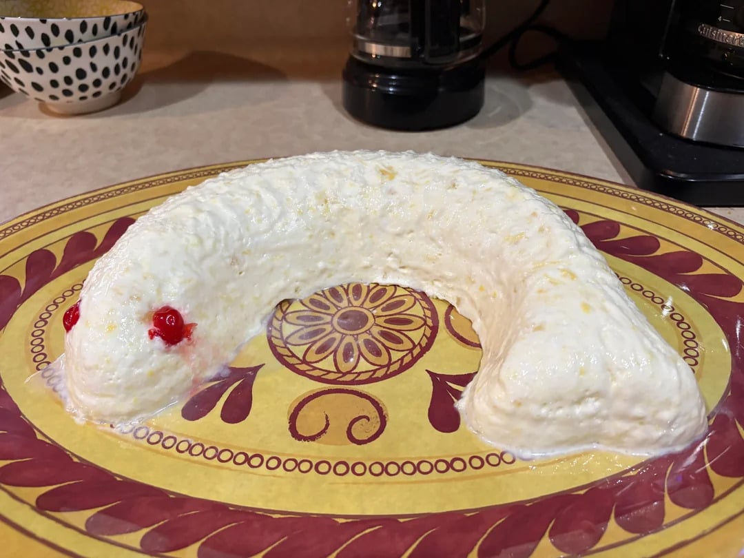 A creamy, white, snake-shaped dessert with two red cherries for eyes sits on a decorative yellow and maroon plate on a kitchen counter.