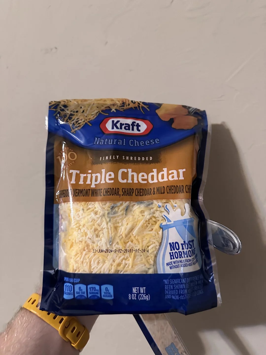 A hand holding a bag of Kraft Natural Cheese Triple Cheddar finely shredded cheese—one of the best shredded cheese options—against a plain wall. The bag is blue and yellow, and the cheese is visible through a clear window on the packaging.
