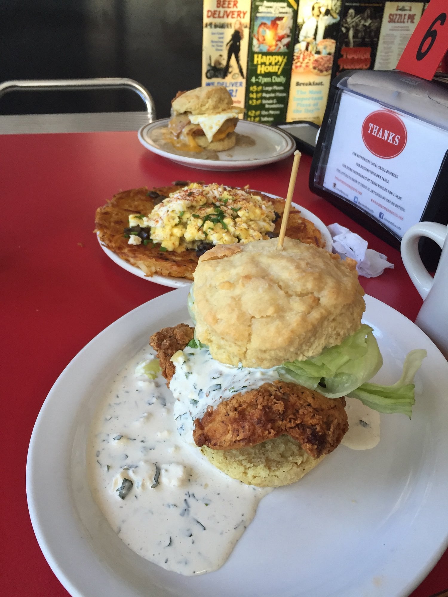 A fried chicken biscuit sandwich with lettuce and creamy sauce on a white plate. In the background are two other breakfast dishes on a red table and a napkin dispenser.