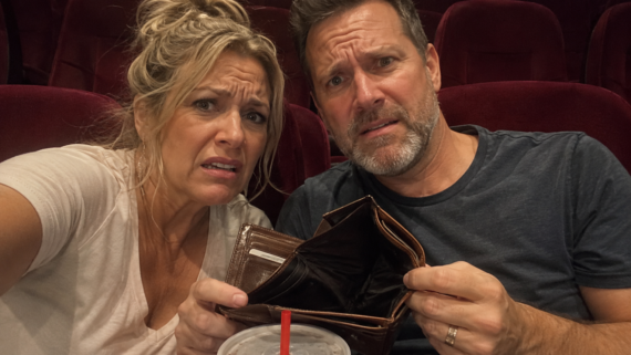 A worried man and woman sit in red theater seats, holding an empty wallet and looking distressed; a drink with a red straw is visible in the foreground, hinting they could use some movie theater hacks to save money.