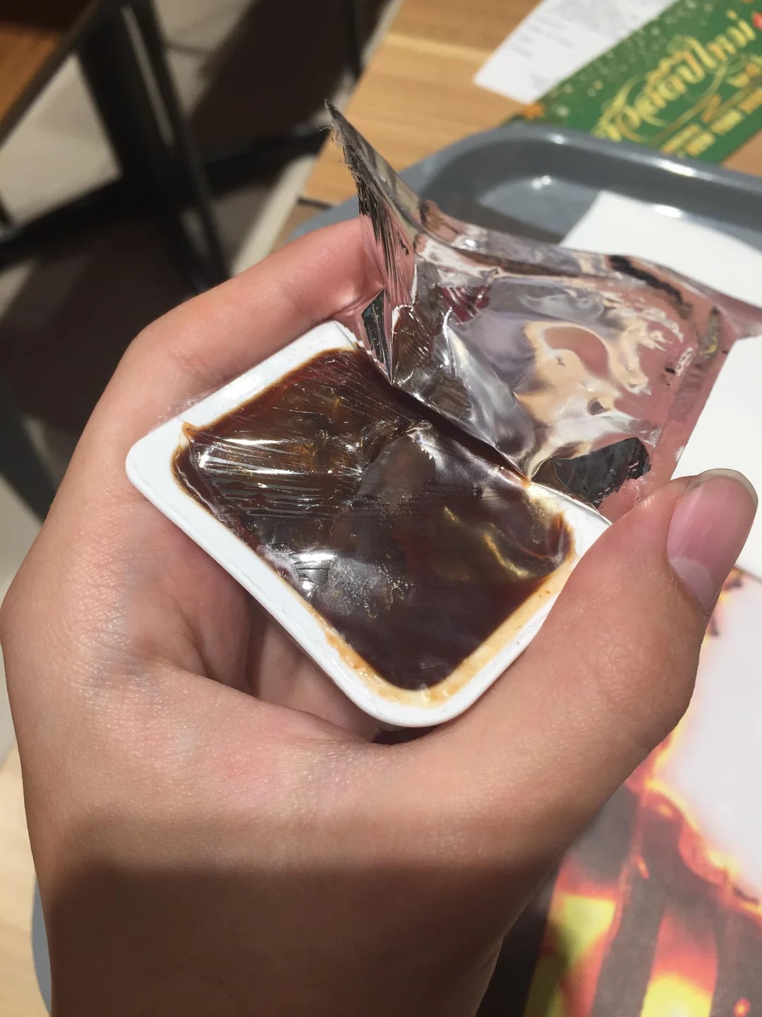 A hand holding an open plastic container of dark brown sauce with the foil lid partially peeled back, over a table with a tray and a menu in the background.