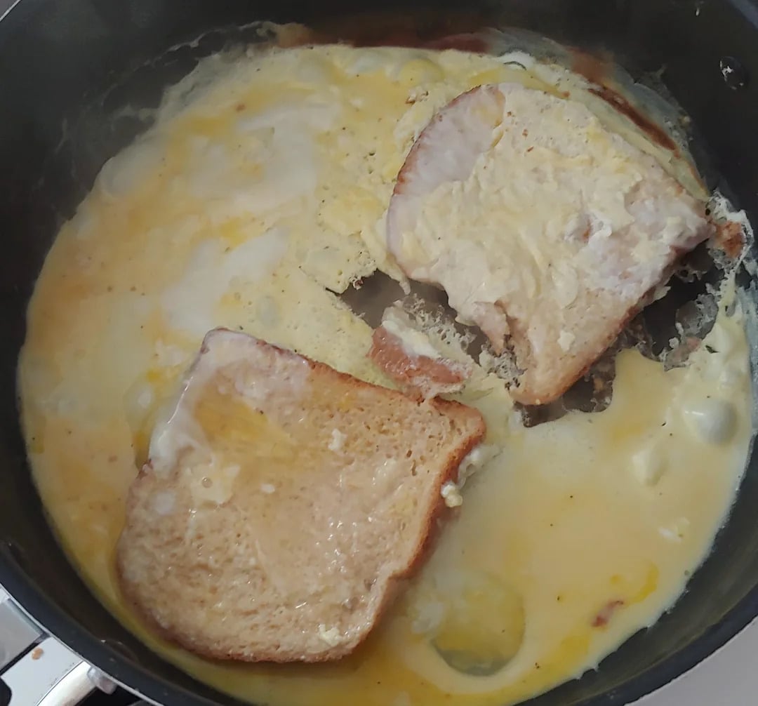 Two slices of bread soaking in beaten eggs cooking in a frying pan, resembling the beginning of making French toast.