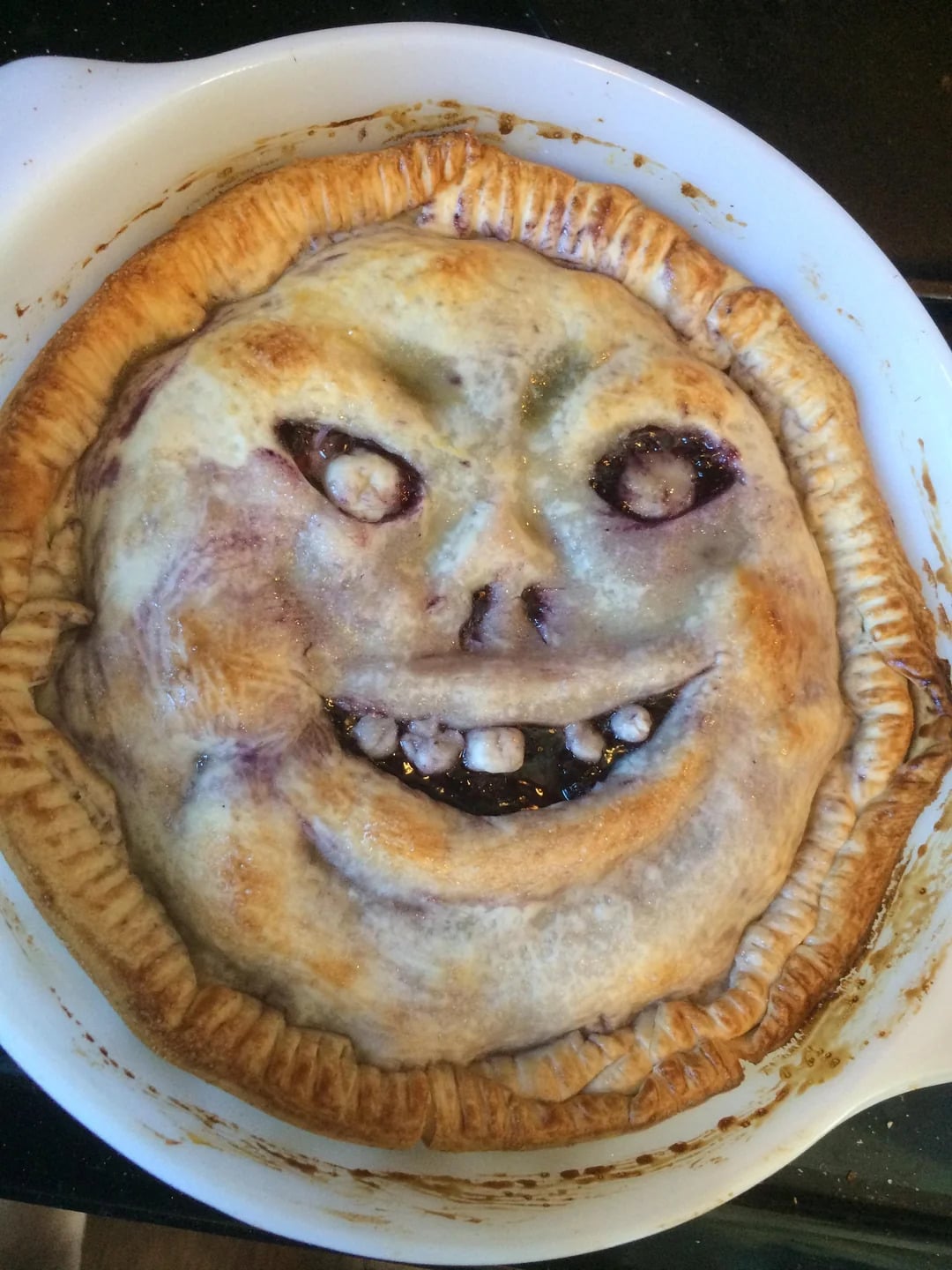 A baked pie with a crust designed to look like a creepy, smiling face with uneven eyes, a nose, and visible teeth, giving it a spooky appearance.