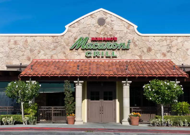The entrance of a Romano's Macaroni Grill restaurant with a tiled roof, stone walls, potted plants, and the restaurant name in green and red signage above the doors.