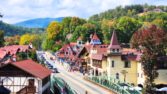 Colorful Bavarian-style tourist town with alpine architecture, a busy main street filled with pedestrians and cars, and green hills in the background under a partly cloudy sky.