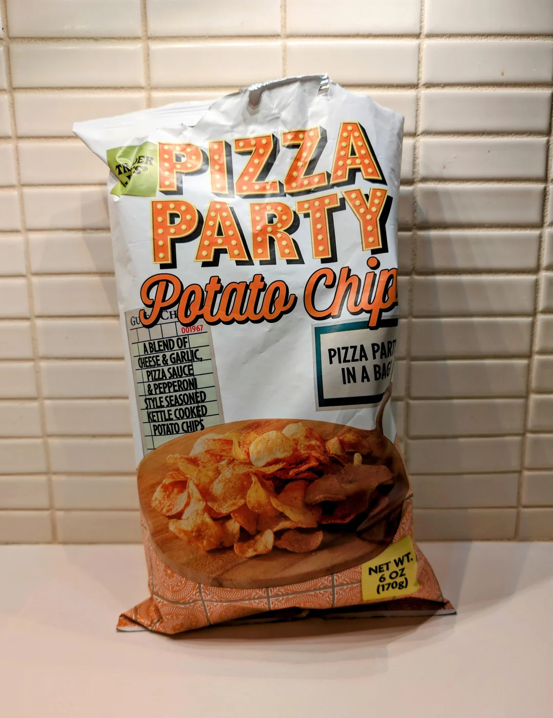 A bag of Pizza Party Potato Chips sits on a counter against a tiled wall. The package has an image of chips, pizza-themed graphics, and lists ingredients like cheese, garlic, and tomato. Net weight is 6 oz (170g). New Trader Joe's products.