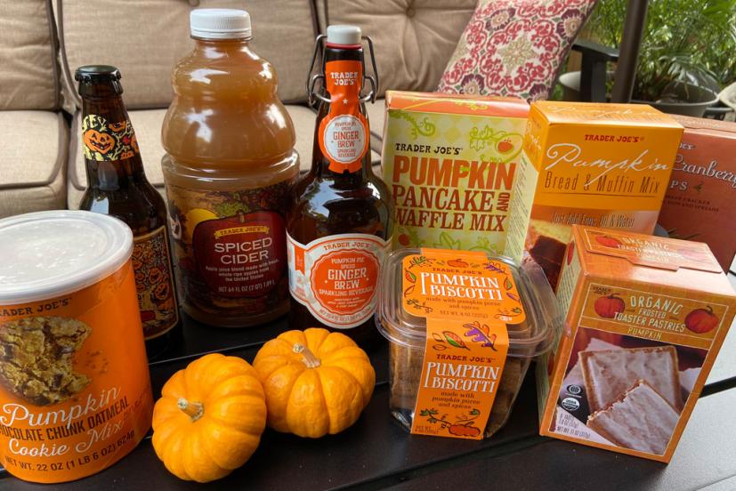 A variety of Trader Joe's pumpkin and fall-themed products, including cookies, pancake mix, cider, ginger brew, biscotti, and pastries, displayed with two small pumpkins on a table outdoors.