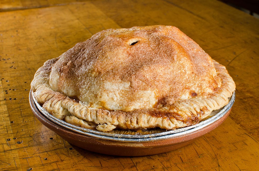 A whole, golden-brown, homemade apple pie with a domed, flaky crust sits in a pie tin on a wooden surface. The crust is sprinkled with sugar and shows slight bubbling at the edges. Classic apple pies never looked so inviting!