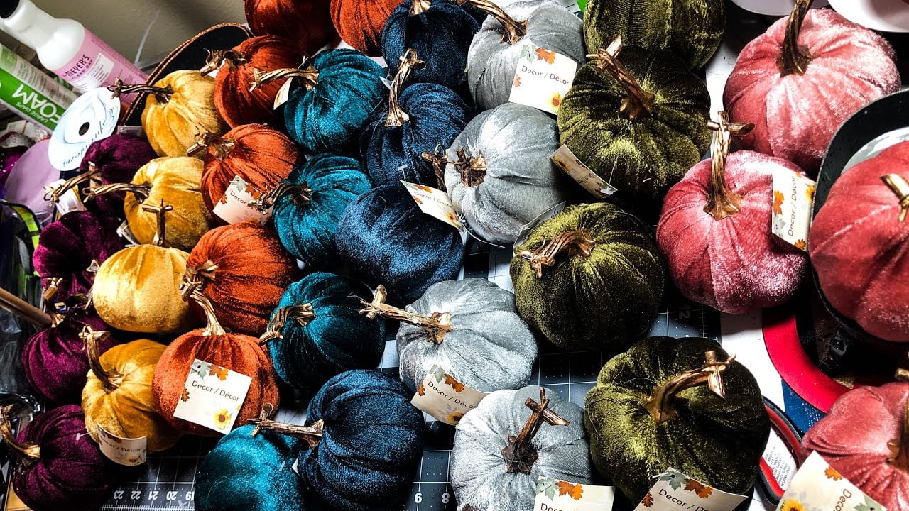 A variety of small, velvet pumpkins in different colors are arranged on a grid surface. Each pumpkin has a brown stem and some have tags attached. Craft supplies are visible in the background.