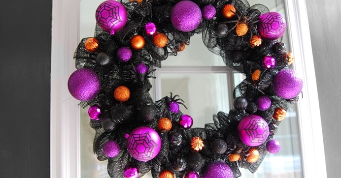 Black Halloween wreath decorated with purple and orange glittery ornaments, some with spiderweb designs, and small black and orange accents, hanging on a window.