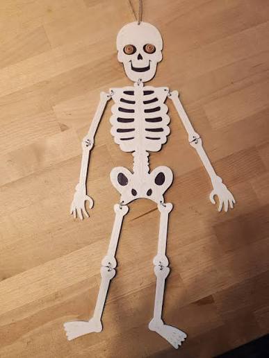 A paper skeleton decoration with movable joints and a string at the top is lying flat on a wooden surface. The skeleton has round button eyes and a simple smiling face.