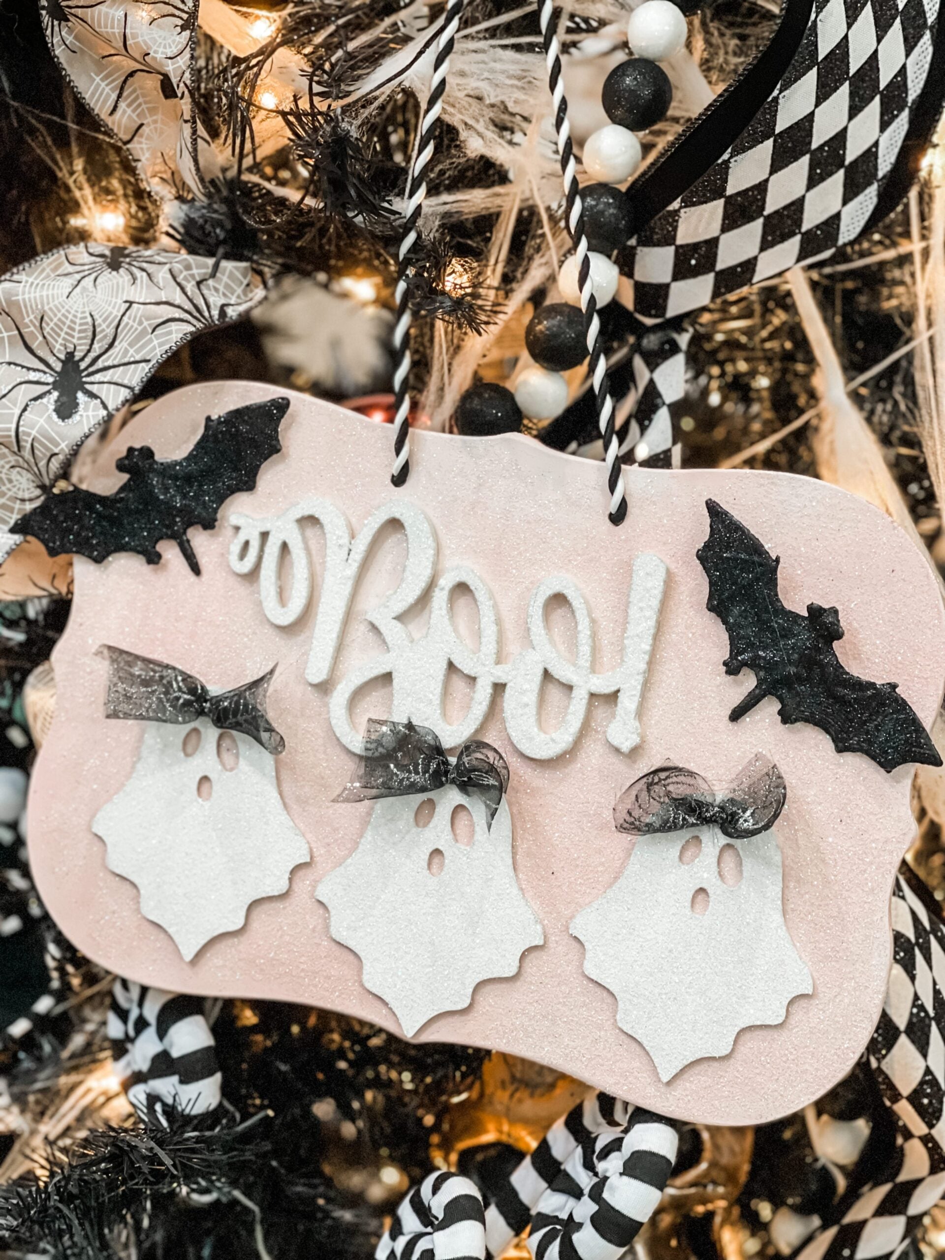 A Halloween decoration with three white ghost figures, the word "Boo!" in white, and two black bats, all hanging on a tree with black-and-white ornaments and cobwebs.