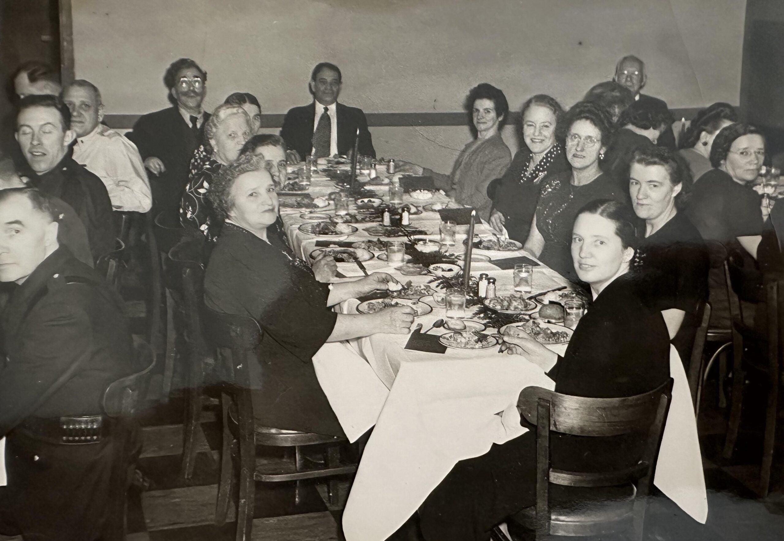 A black-and-white photo of about 17 people sitting around a long dinner table, eating and smiling, dressed in formal attire, in a restaurant or banquet setting—capturing the timeless warmth of nostalgic dinners shared among friends and family.