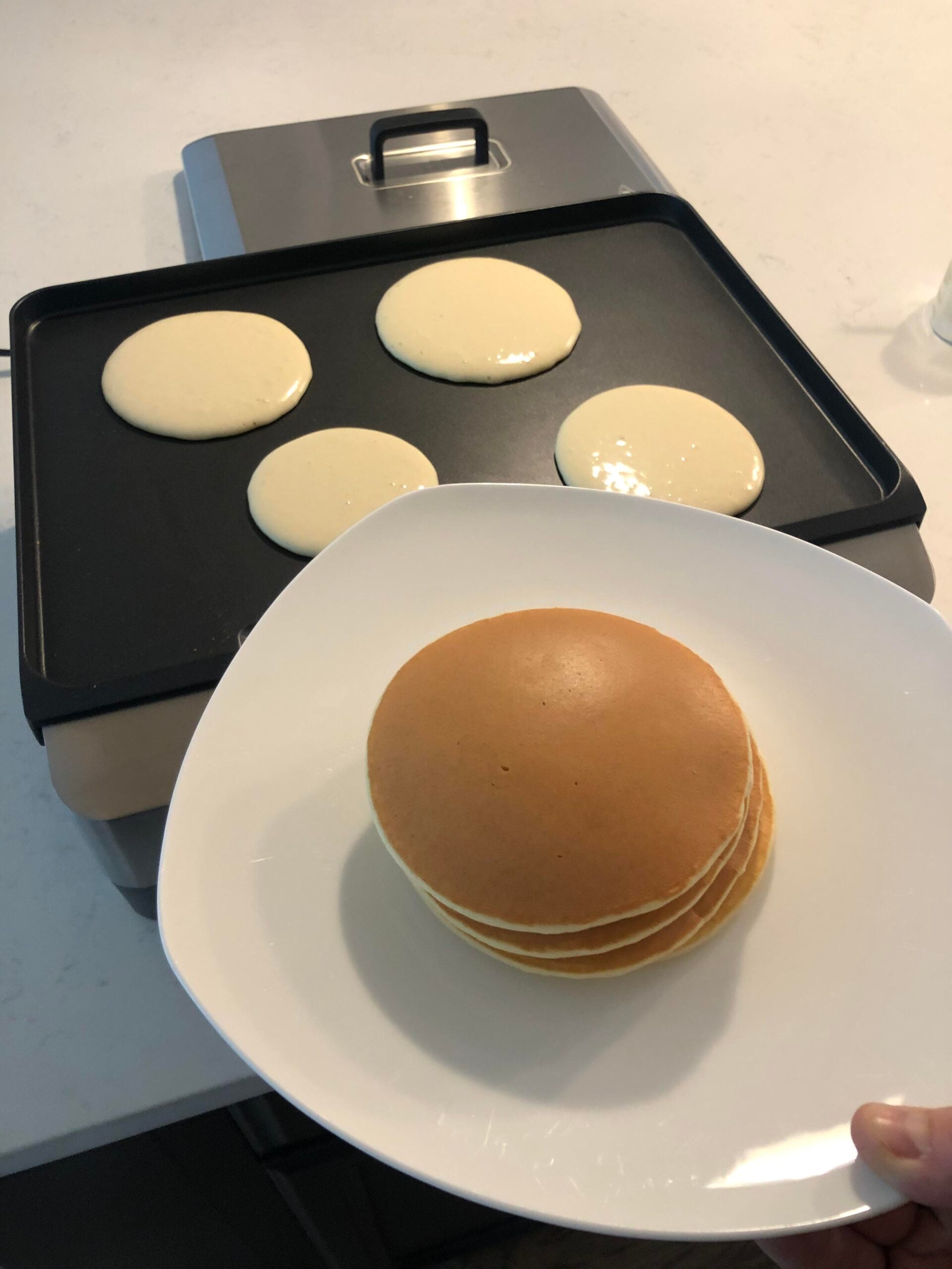 A hand holds a plate with a stack of cooked pancakes, while four uncooked pancakes are cooking on an electric griddle in the background.