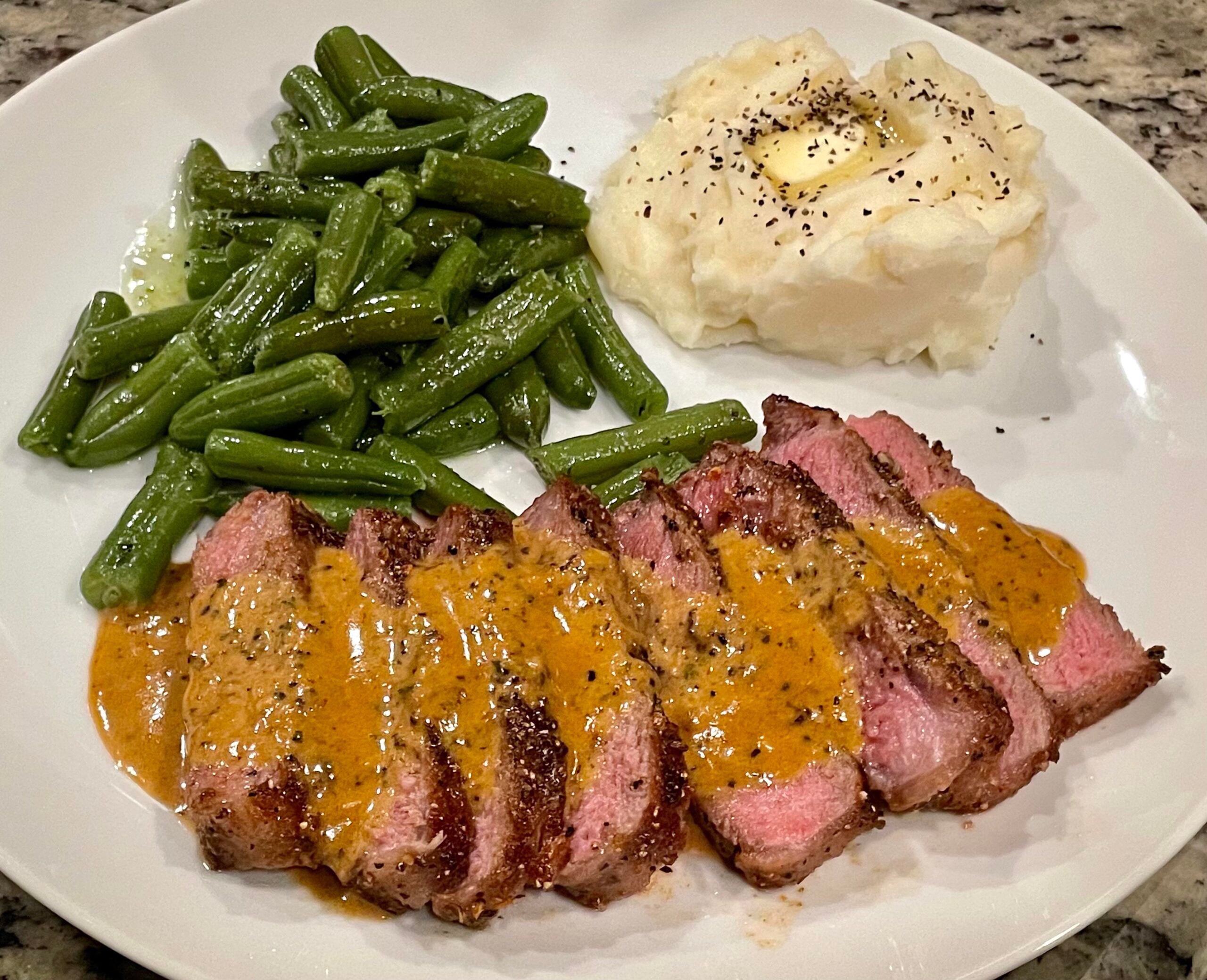 Sliced steak with creamy orange sauce, served with green beans and mashed potatoes topped with butter and black pepper on a white plate.