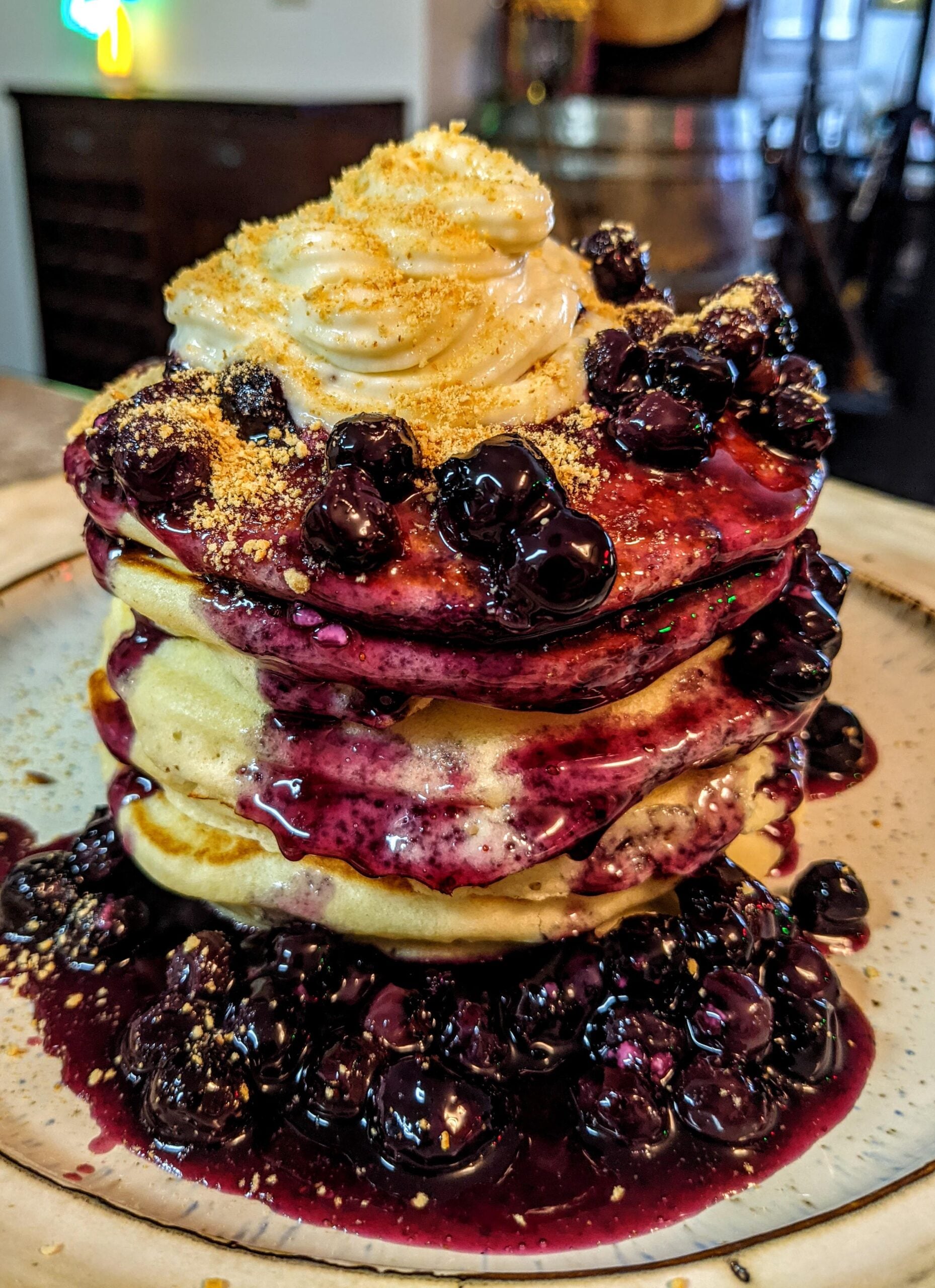 A stack of homemade pancakes topped with whipped cream, blueberry compote, and a sprinkle of brown sugar, served on a ceramic plate.