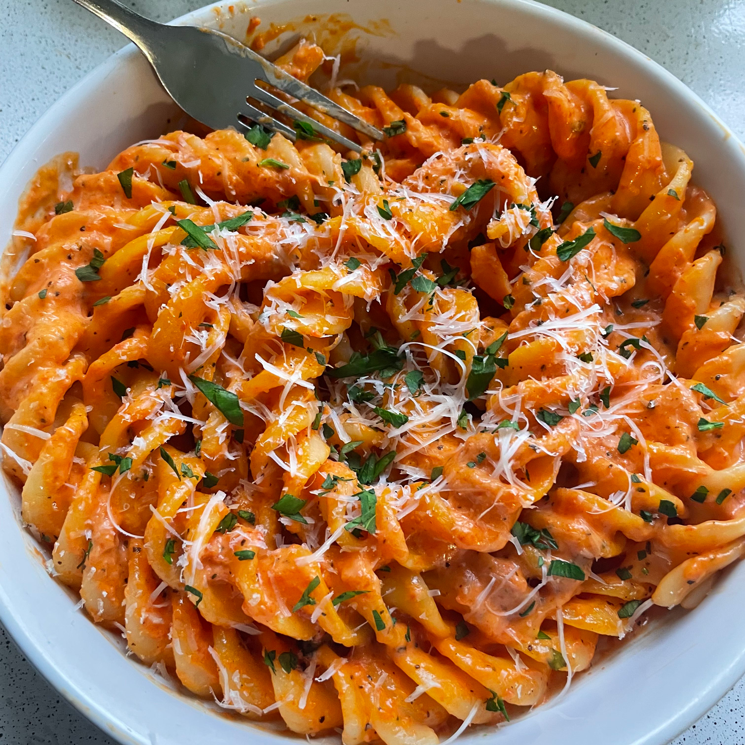 A bowl of rotini pasta covered in creamy tomato sauce, topped with grated cheese and chopped herbs, with a fork resting on the side of the bowl.