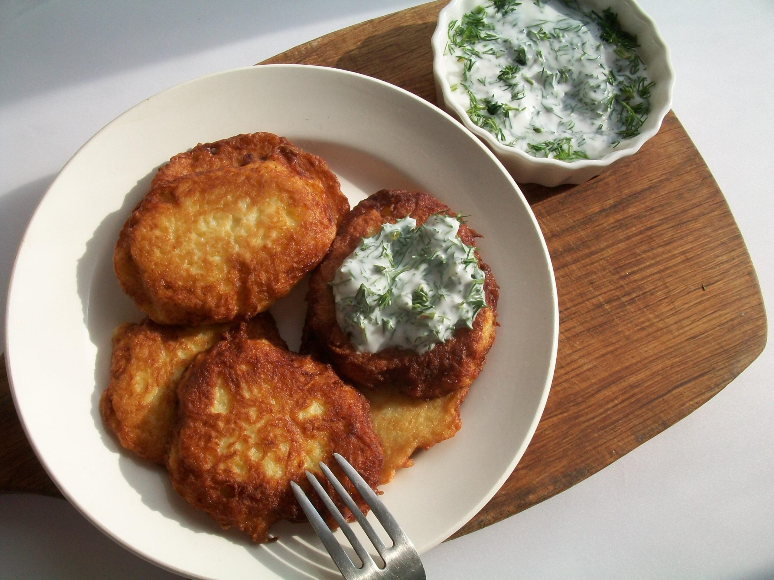 Four golden-brown homemade potato pancakes rest on a white plate, one topped with creamy dill sauce, with a fork beside them. A small bowl of sauce sits nearby on a wooden board.